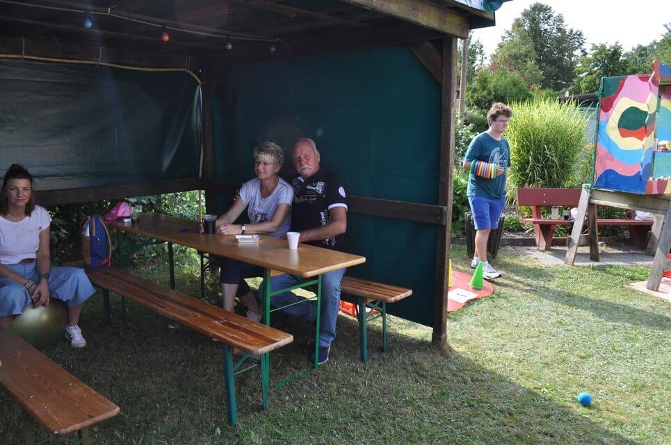Kinderfest im Kleingartenverein Gäste sitzen auf Bänken in einem schattigen Gartenunterstand.